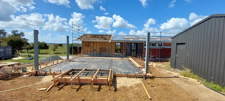 New house slab boxed and poured, with the timber-framed shell going up on it beside a farm shed
